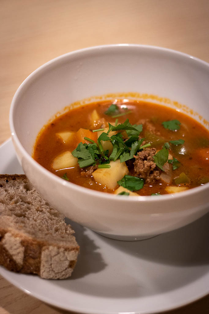 Dyp skål med ferdig gulasjsuppe toppet med hakket bladpersille og en skive brød ved siden av