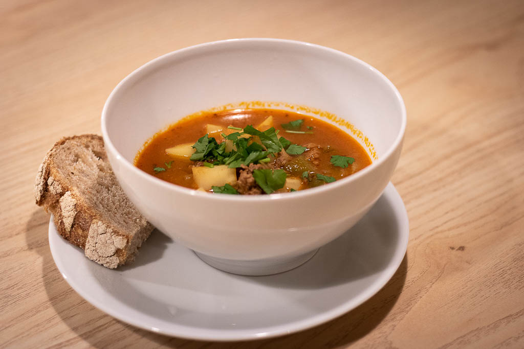 Dyp skål med ferdig gulasjsuppe toppet med hakket bladpersille og en skive brød ved siden av